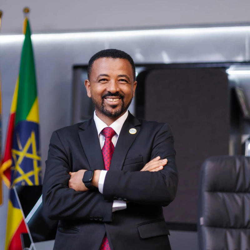 Dr. Asrat Atsedeweyn standing with folded arms, wearing a dark suit and red tie